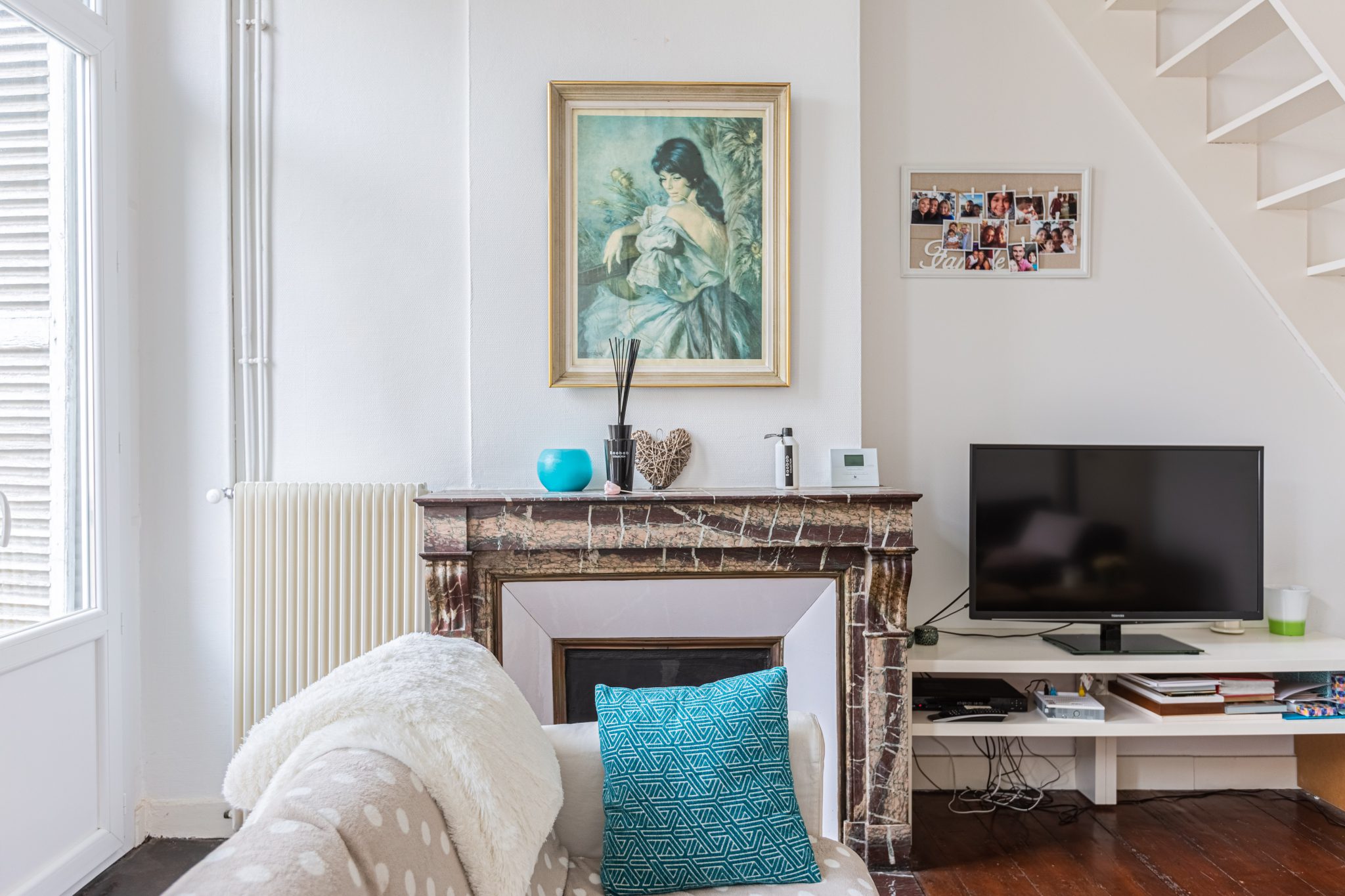 petit salon lumineux avec une cheminée, un coussin bleu, un tableau d'art et une télé